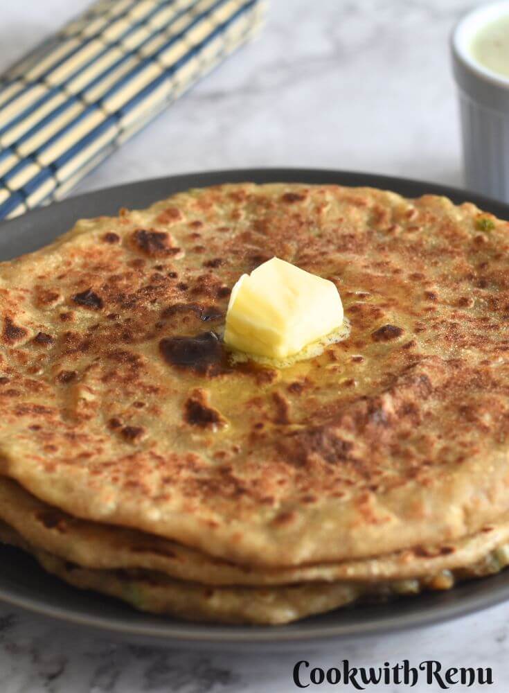 Aloo Paratha in a grey plate, with a dollop of butter. Seen in the background is pudina raita.