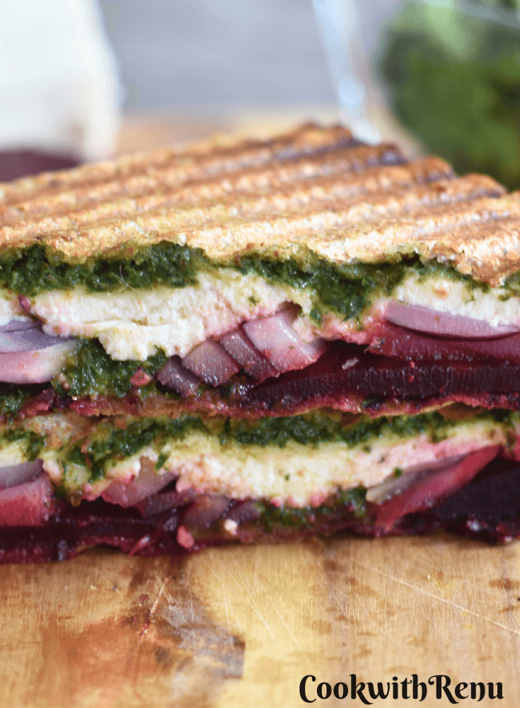 Close up look of Carrot Greens Pesto Beetroot Paneer Sandwich, which is cut open. Seen are the layers of pesto, paneer, onion and beetroot. Seen in the background is Paneer, Beetroot slices and Carrot greens pesto.