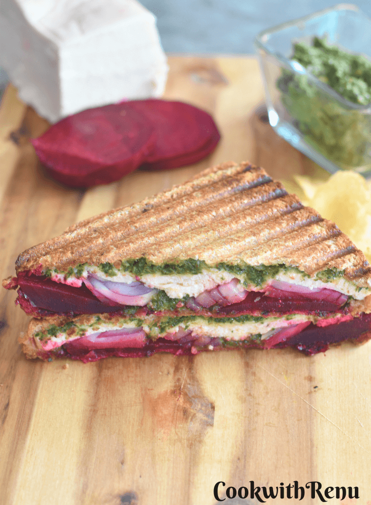 Top and close up look of Carrot Greens Pesto Beetroot Paneer Sandwich, which is cut open. Seen are the layers of pesto, paneer, onion and beetroot. Seen in the background is Paneer, Beetroot slices and Carrot greens pesto.