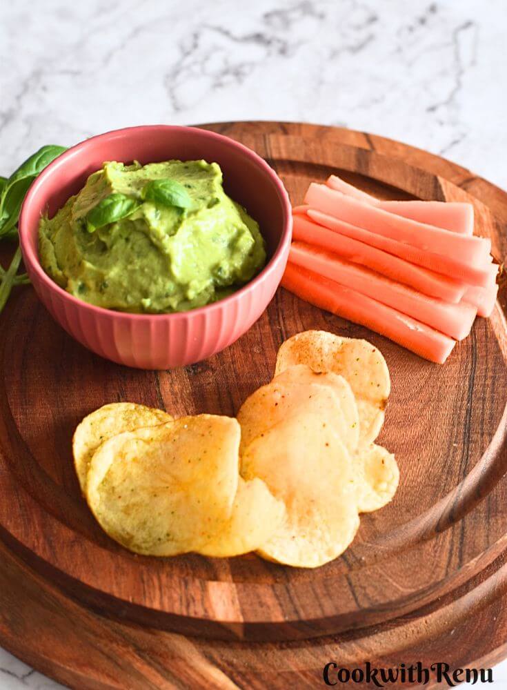 Avocado Basil Dip served with chips and carrot on a brown cheese board.