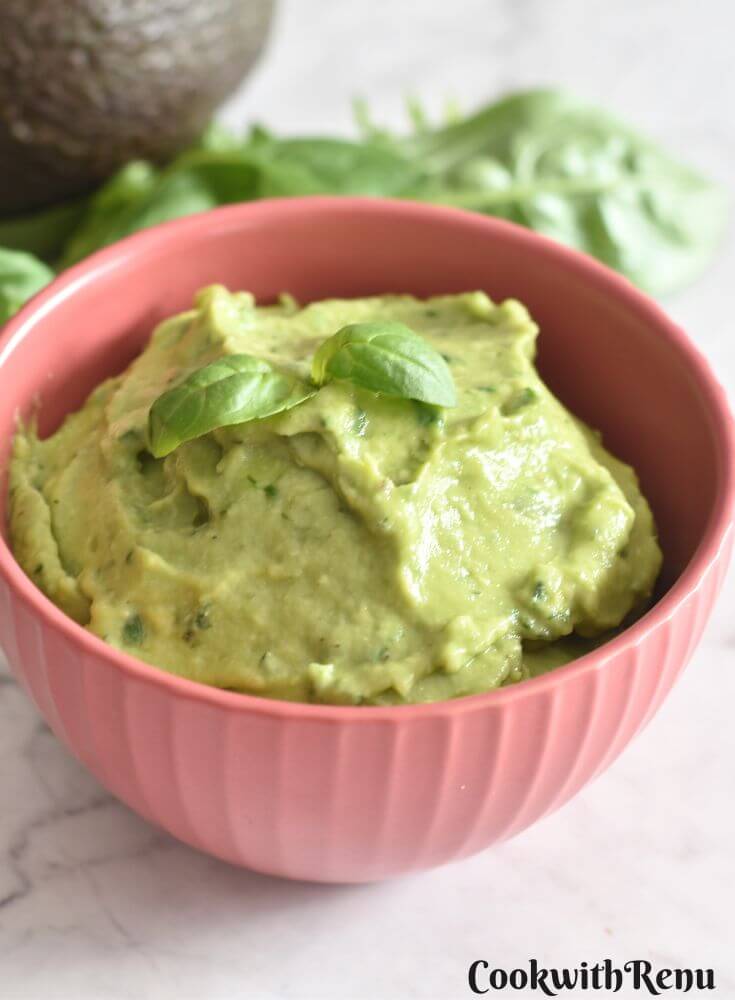 Avocado Basil Dip served in a red bowl, with a garnish of basil on top. Seen in the background are some basil leaves and avocado.