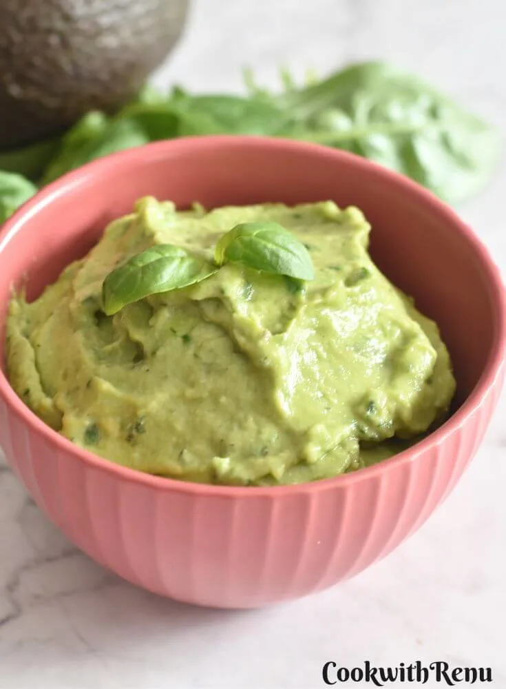 Avocado Basil Dip served in a red bowl, with a garnish of basil on top. Seen in the background are some basil leaves and avocado.