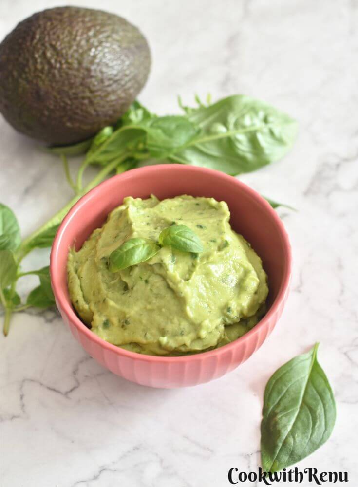 Avocado Basil Dip served in a red bowl, with a garnish of basil on top. Seen in the background are some basil leaves and avocado.