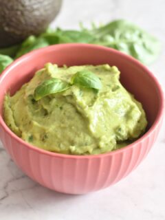 Avocado Basil Dip served in a red bowl, with a garnish of basil on top. Seen in the background are some basil leaves and avocado.