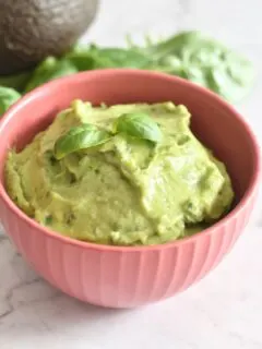 Avocado Basil Dip served in a red bowl, with a garnish of basil on top. Seen in the background are some basil leaves and avocado.