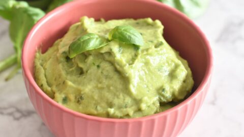 Avocado Basil Dip served in a red bowl, with a garnish of basil on top. Seen in the background are some basil leaves and avocado.
