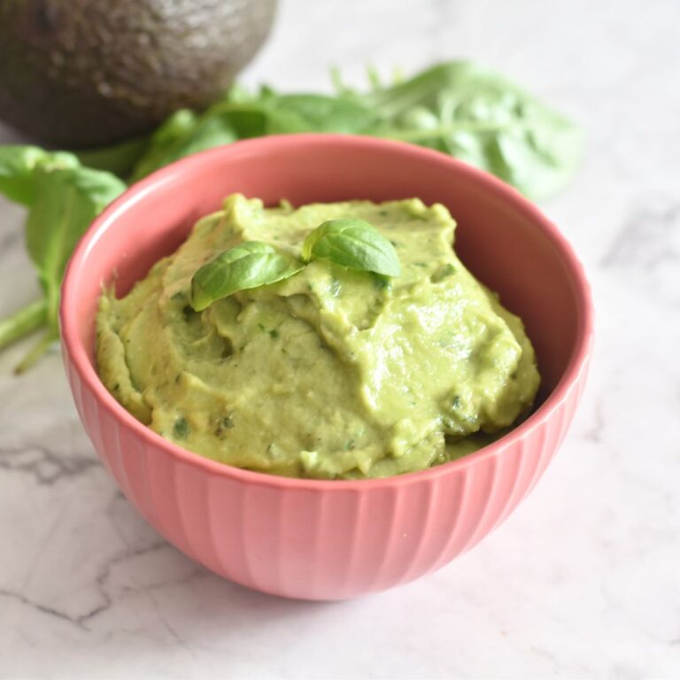 Avocado Basil Dip served in a red bowl, with a garnish of basil on top. Seen in the background are some basil leaves and avocado.