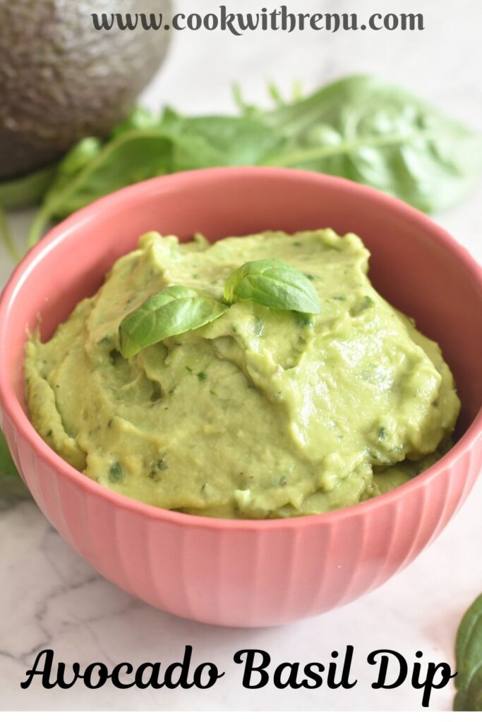 Avocado Basil Dip served in a red bowl, with a garnish of basil on top. Seen in the background are some basil leaves and avocado.
