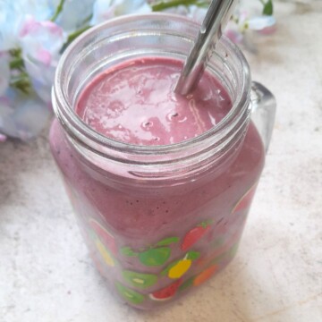 Banana Berry Protein Smoothie served in a glass with a steel straw in it. Seen in the background are some flowers.