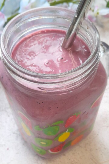 Banana Berry Protein Smoothie served in a glass with a steel straw in it. Seen in the background are some flowers.