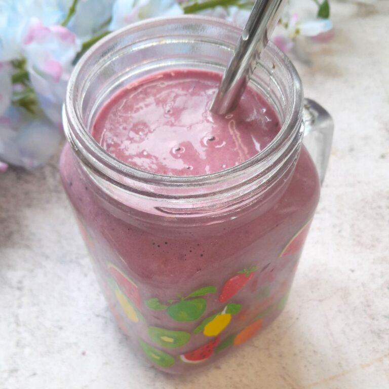 Banana Berry Protein Smoothie served in a glass with a steel straw in it. Seen in the background are some flowers.