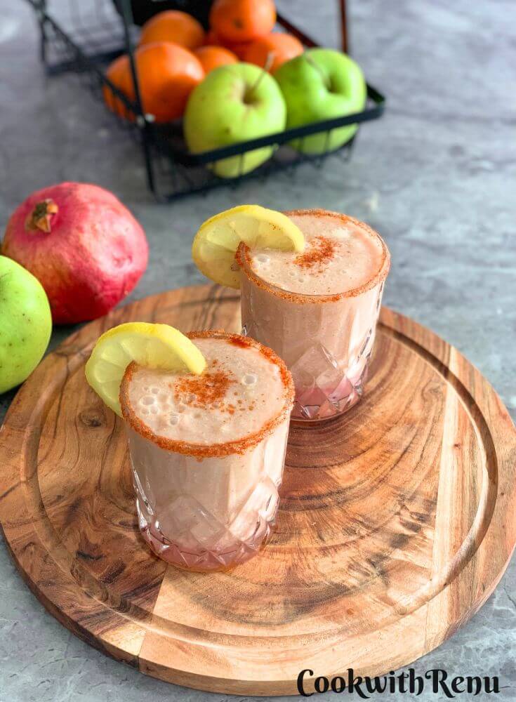 Apple Pomegranate Juice served in 2 glasses with a garnish of lemon and some salt, and spice mix. Seen in the background are some fruits.