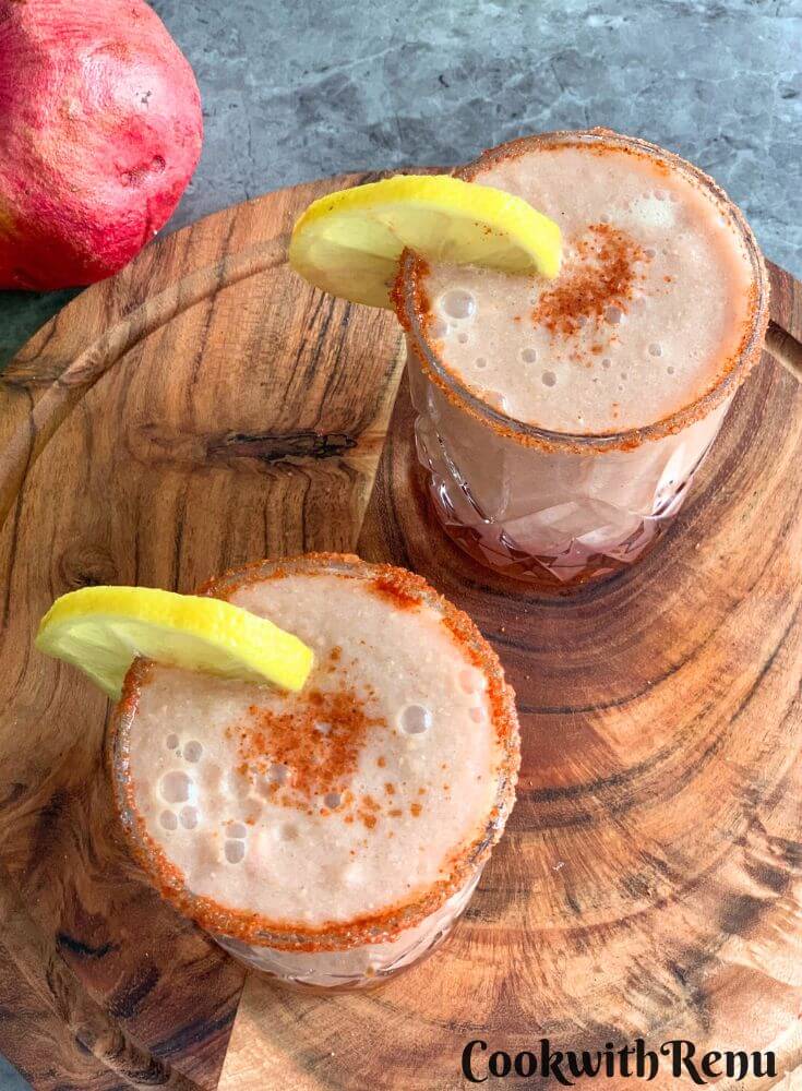 Apple Pomegranate Juice served in 2 glasses with a garnish of lemon and some salt, and spice mix. Seen in the background are some fruits.