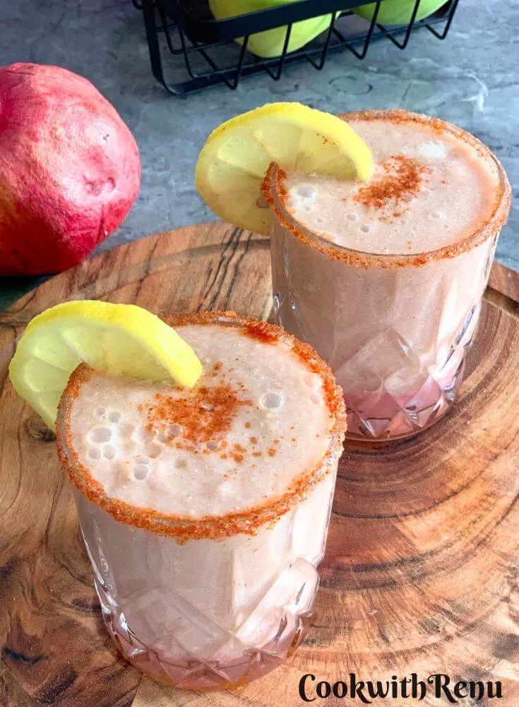 Apple Pomegranate Juice served in 2 glasses with a garnish of lemon and some salt, and spice mix. Seen in the background are some fruits.