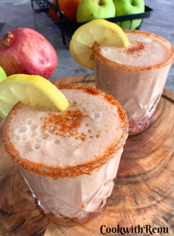Apple Pomegranate Juice served in 2 glasses with a garnish of lemon and some salt, and spice mix. Seen in the background are some fruits.