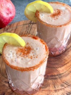 Apple Pomegranate Juice served in 2 glasses with a garnish of lemon and some salt, and spice mix. Seen in the background are some fruits.