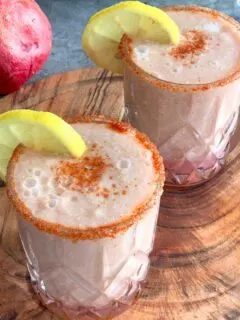 Apple Pomegranate Juice served in 2 glasses with a garnish of lemon and some salt, and spice mix. Seen in the background are some fruits.