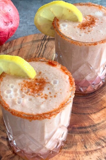 Apple Pomegranate Juice served in 2 glasses with a garnish of lemon and some salt, and spice mix. Seen in the background are some fruits.