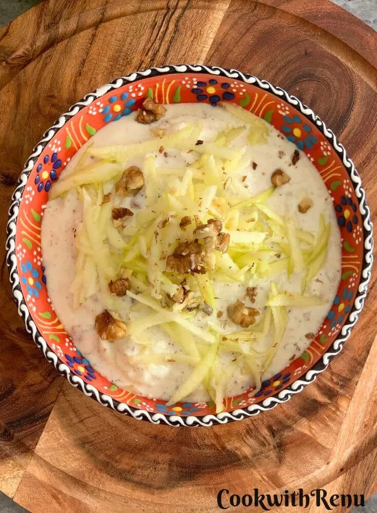 Top view of Green Apple Walnut Raita served in an orange designer bowl. Seen in the background are some crisp green apples and a tree.