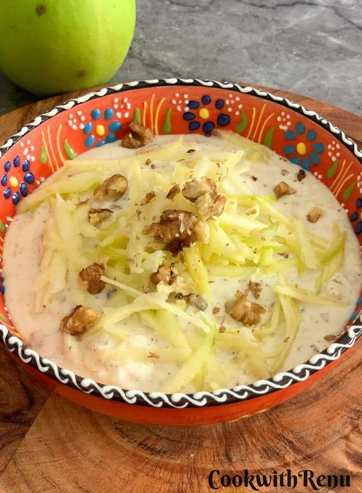 Green Apple Walnut Raita served in an orange designer bowl. Seen in the background are some crisp green apples and a tree.