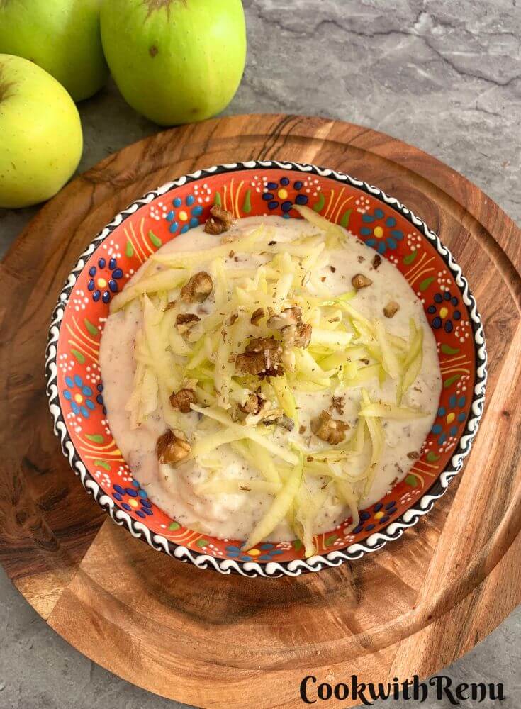 Top view of Green Apple Walnut Raita served in an orange designer bowl. Seen in the background are some crisp green apples and a tree.