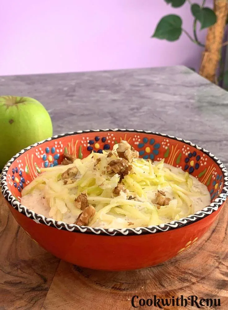Green Apple Walnut Raita served in an orange designer bowl. Seen in the background are some crisp green apples and a tree.