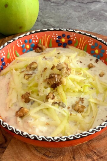Green Apple Walnut Raita served in an orange designer bowl. Seen in the background are some crisp green apples and a tree.