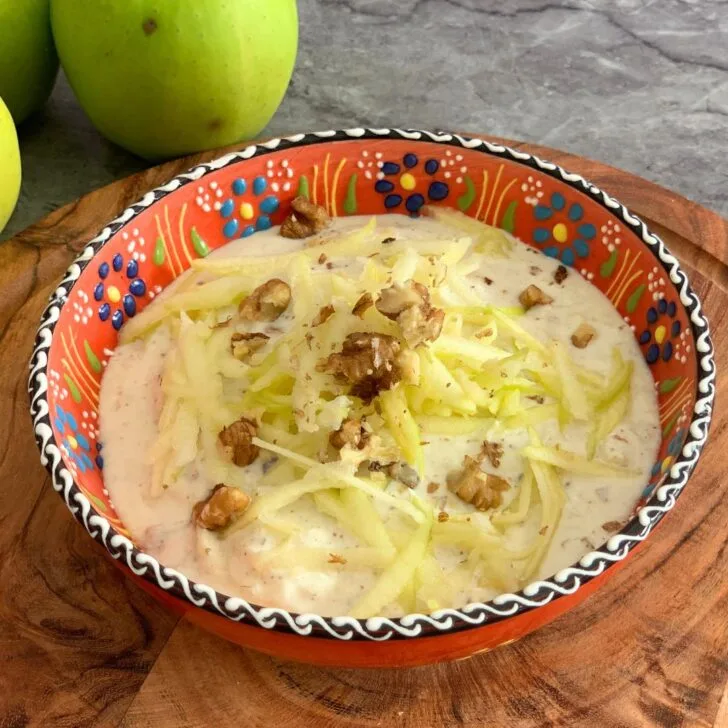 Green Apple Walnut Raita served in an orange designer bowl. Seen in the background are some crisp green apples and a tree.