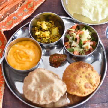 Image showing an Indian Thali served in a steel plate with steel bowls. In the image, from 6 O Clock and clock wise, Sada Puri, aamras in a bowl, aloo bhaji in a bowl, salad in a bowl, missi puri, and homemade mango pickle in the center are served along with papad in another small steel plate.