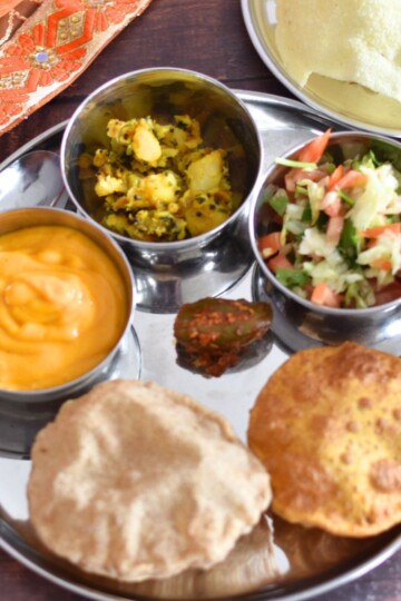 Image showing an Indian Thali served in a steel plate with steel bowls. In the image, from 6 O Clock and clock wise, Sada Puri, aamras in a bowl, aloo bhaji in a bowl, salad in a bowl, missi puri, and homemade mango pickle in the center are served along with papad in another small steel plate.