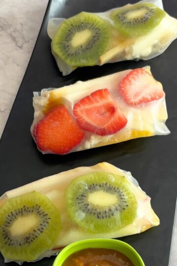 Three Fruit Summer Rolls served on a black tray and some mango chili sauce as a dip.