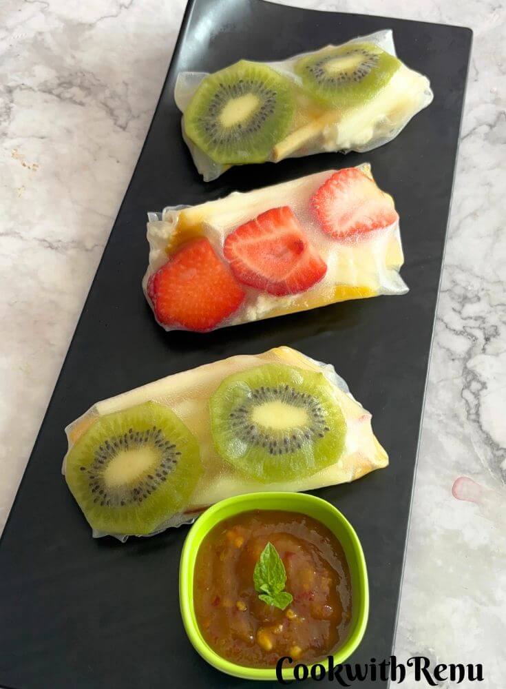 Three Fruit Summer Rolls served on a black tray and some mango chili sauce as a dip.