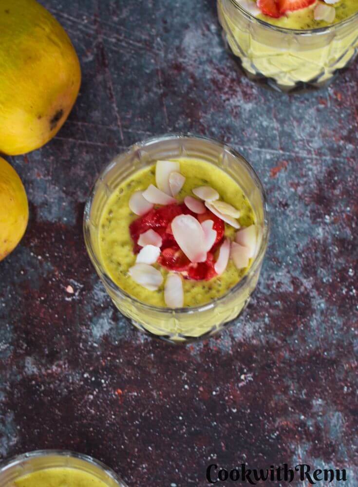 Vegan Mango Chia Seed Pudding served in 3 glasses with a garnish or nuts and strawberry pieces. Seen in the background are some mangoes.
