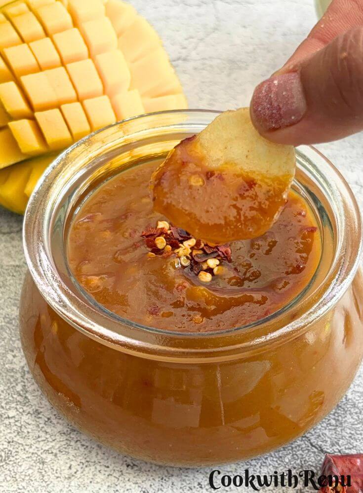 A crisp being dipped in Sweet Mango Chili Sauce in a glass bowl with some chili flakes as garnish, and seen in the background is cut mango.