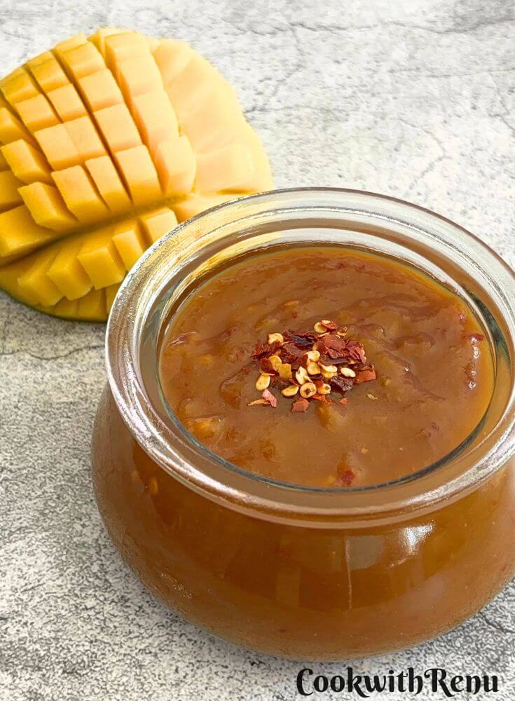 Sweet Mango Chili Sauce Using Jaggery in a glass bowl with some chili flakes as garnish , and seen in the background is cut mango.