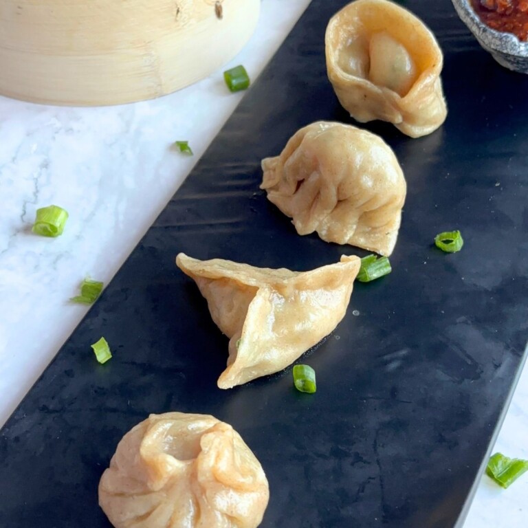 Veg Momos are arranged on a black tray along with spicy garlic chutney. Seen in the background is momos basket with momos in it.