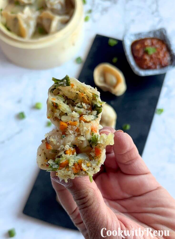 Veg Momos are arranged on a black tray along with spicy garlic chutney. One momo is cut open showing inside stuffing.
