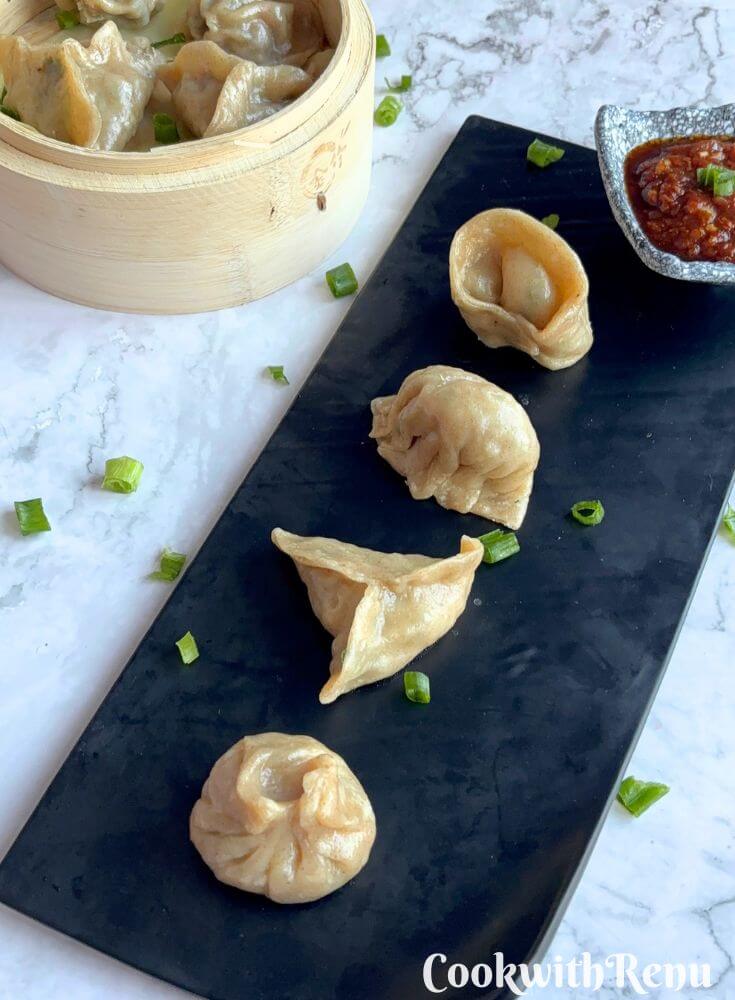 Veg Momos are arranged on a black tray along with spicy garlic chutney. Seen in the background is momos basket with momos in it.