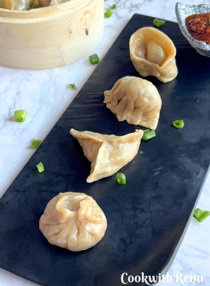 Veg Momos are arranged on a black tray along with spicy garlic chutney. Seen in the background is momos basket with momos in it.