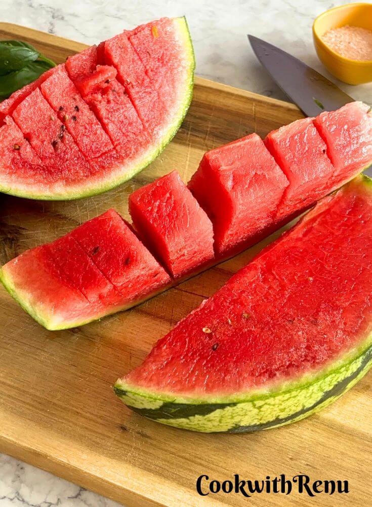 Cut watermelon is seen on a wooden chopping board with some Himalayan salt seen in the background.
