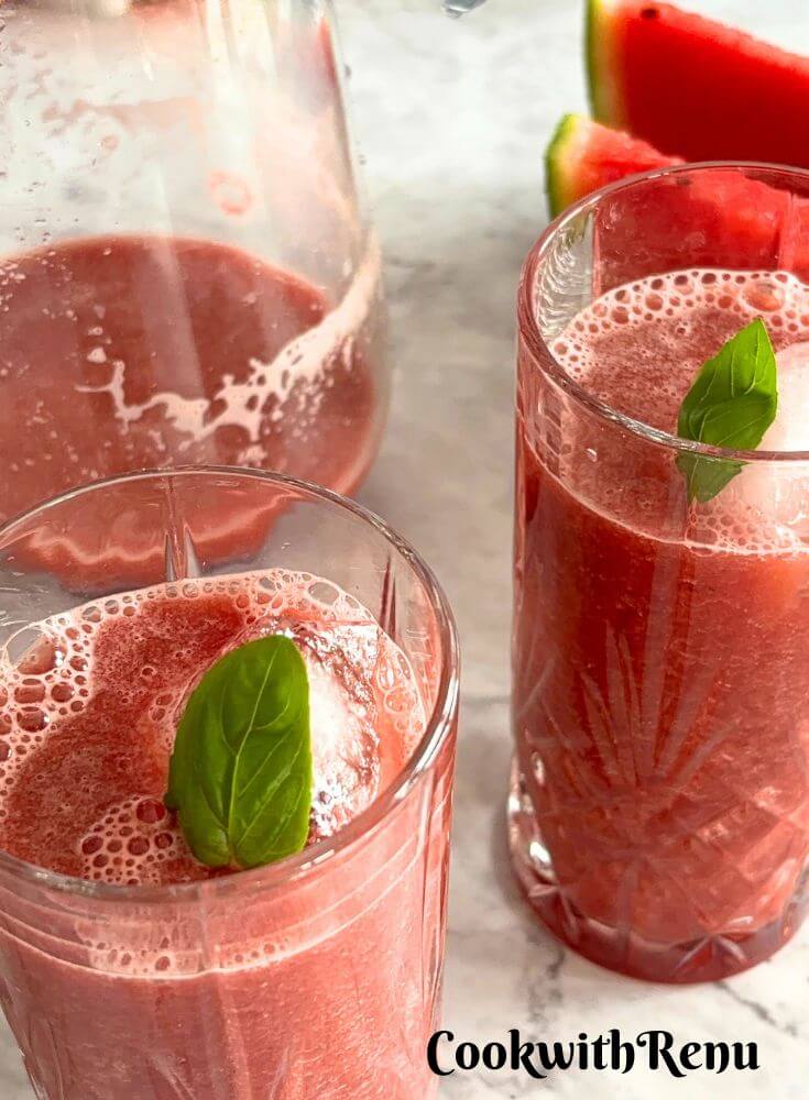 Watermelon Pomegranate Juice served in 2 glasses. In the background is a jug of juice, and seen in the background are 2 slices of watermelon.
