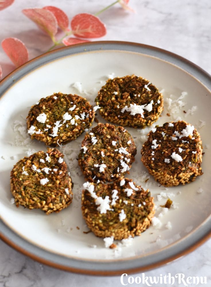 Crispy Maharashtrian Kothimbir Vadi are served in a cream plate with a grey lining. It has a garnish of fresh grated coconut and some artificial flowers in the background.