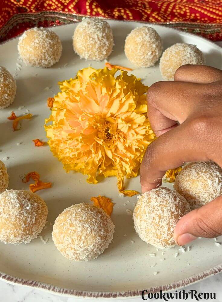 Ladoos are arranged on a cream plate, with a hand seeming to pick one of the ladoos. In the middle are some marigold flowers.