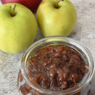 A jar of chunky jaggery apple sauce beside two green apples and one red apple.