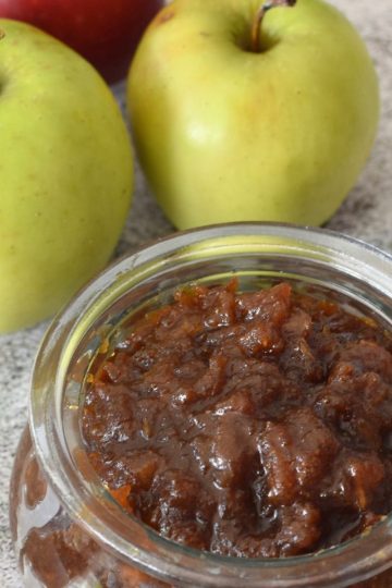A jar of chunky jaggery apple sauce beside two green apples and one red apple.